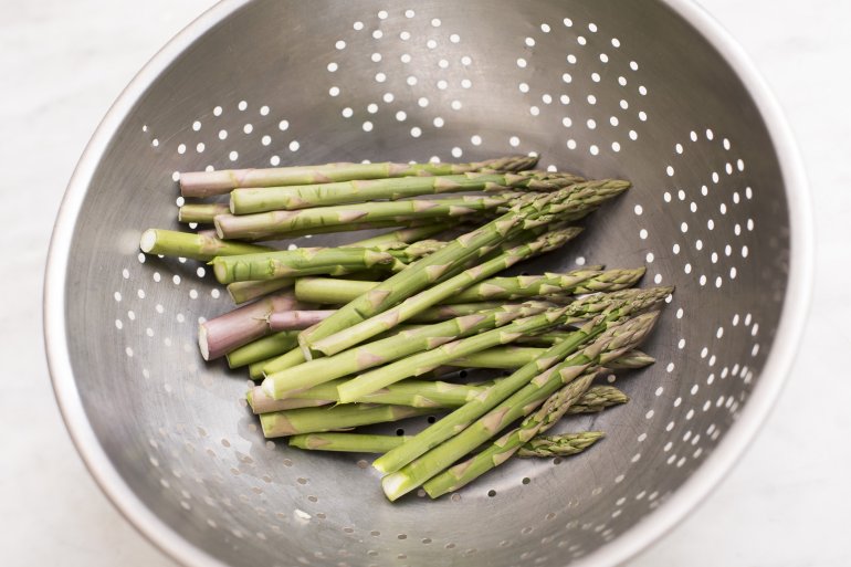 Cut raw asparagus spears sitting in middle of large metal colander with little holes to drain water