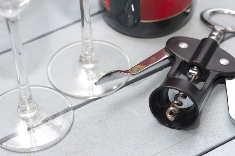 Lever style corkscrew bottle opener alongside wineglasses and a bottle of wine on a white wooden table