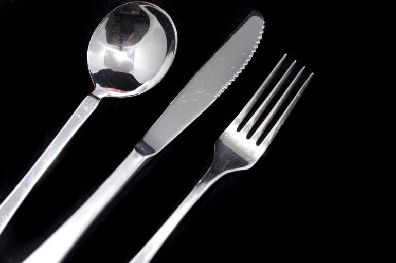 Clean metal knife, fork and spoon on black background with copy space arranged diagonally from the left corner