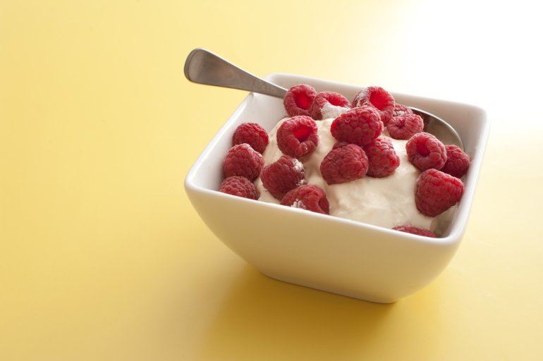 Delicious creamy yoghurt topped with fresh red raspberries served in a modern square bowl over a yellow background with copy space