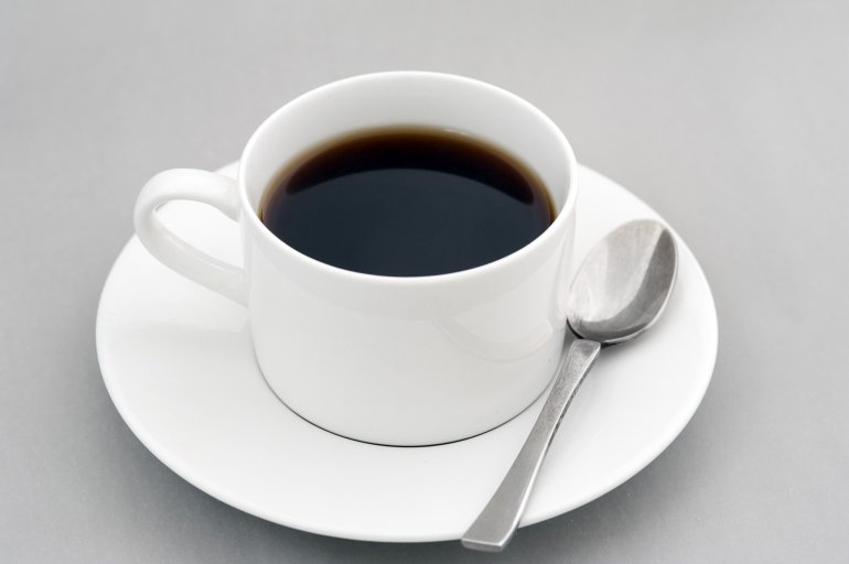 Cup of strong black espresso coffee served in a generic white ceramic cup and saucer with teaspoon over a grey background