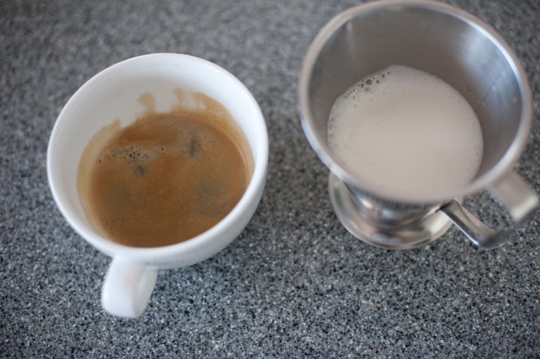 Cup of strong espresso coffee with frothy heated milk alongside in a metal container from a coffee machine, high angle view on a dark grey background