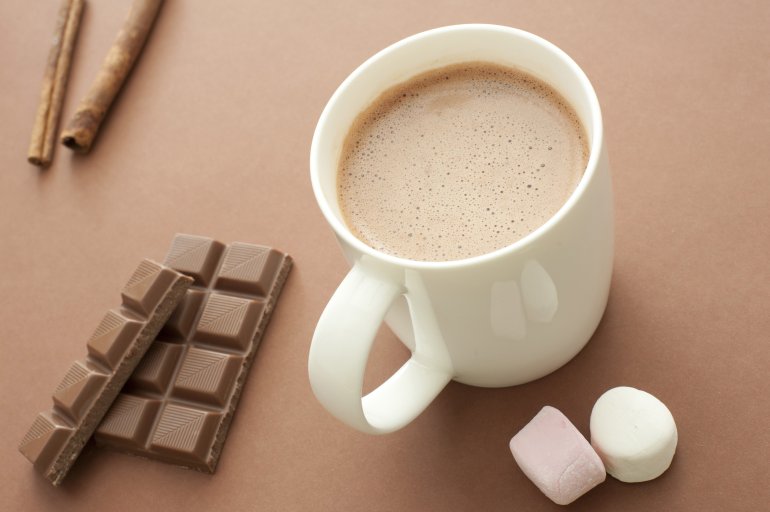 Delicious hot chocolate drink in a mug surrounded by chocolate candy, marshmallows and aromatic stick cinnamon on a brown background, high angle view