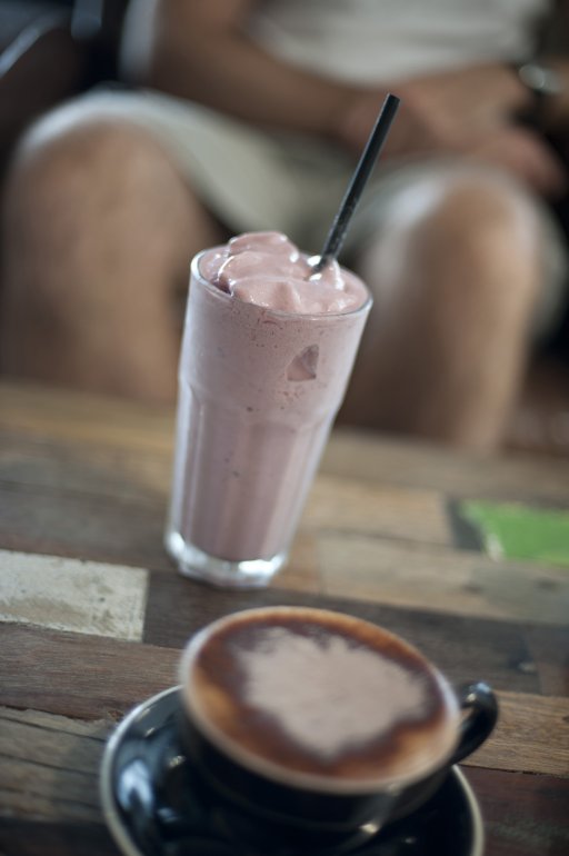 Fruit milkshake standing alongside a cup of cappuccino on a restaurant table, legs of a person seated at the table visible in the background