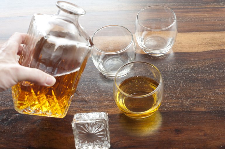 Man pouring whiskey from a glass decanter into glasses or tumblers on a wooden bar counter, close up high angle view of his hand