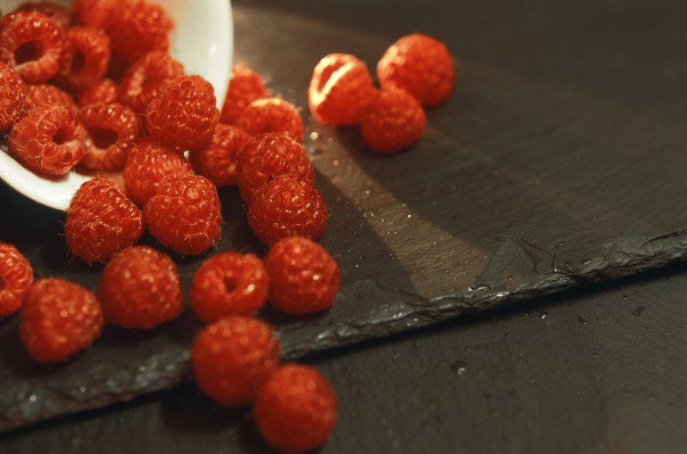Ripe red autumn raspberries spilling out of a ceramic dish onto an old wooden tabletop, closeup view