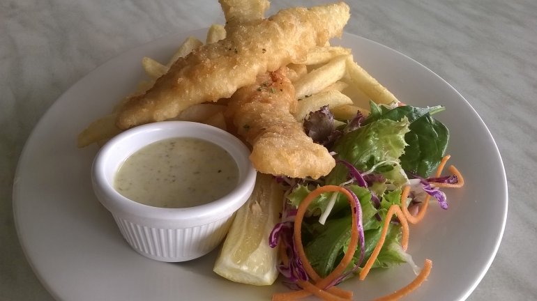Battered fish and fried potato chips served with a savory sauce and a fresh salad on a white plate, close up high angle view