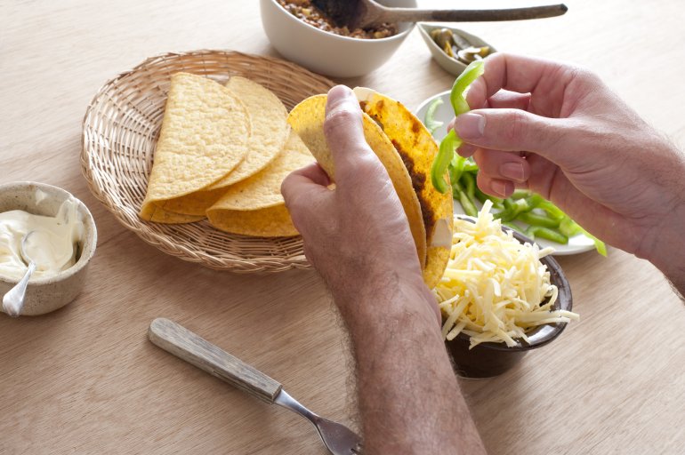 First person perspective view of man making hard shelled tacos using ground beef and shredded cheese beside small container of sour cream and sliced green pepper