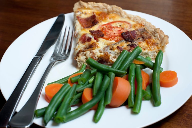 Slice of savoury quiche with fresh cooked carrots and green beans served on a white plate with cutlery over a black background