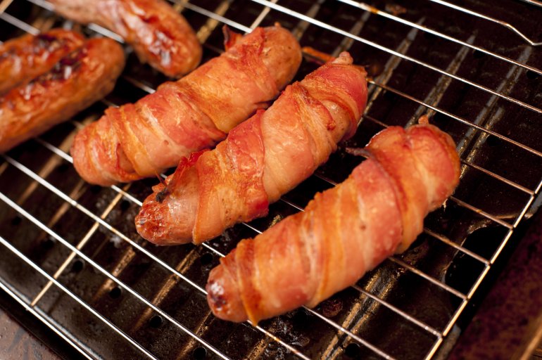 Bacon rolls or pigs in blankets grilling on a BBQ with pork sausages wrapped in rashers of bacon in close up on the grid