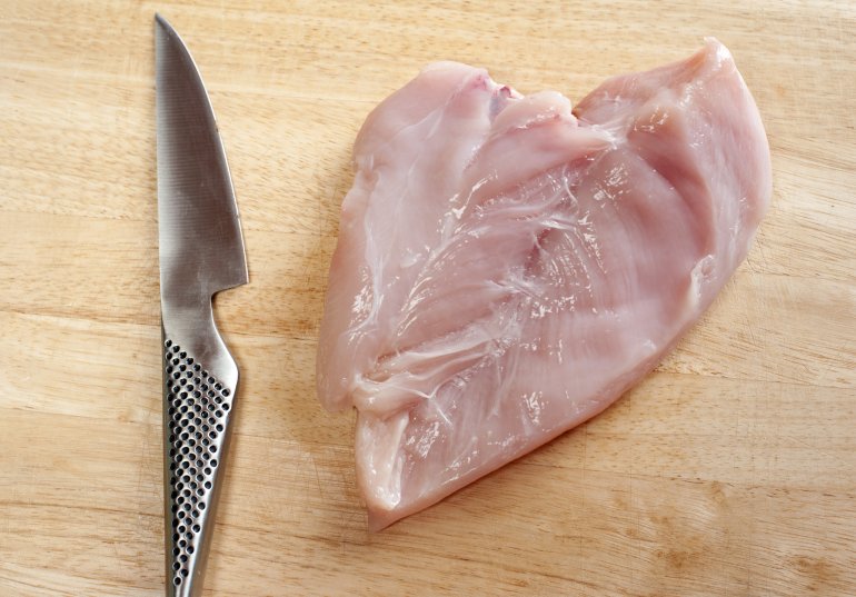 Raw chicken breast cut in a butterfly on a wooden chopping board with a sharp kitchen knife alongside, overhead view