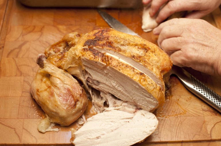Whole roast chicken sliced through the breast with a mans hands and carving knife visible at the side, high angle view