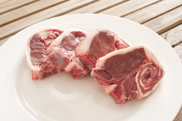 Four fresh raw lamb cutlets with a fatty rind displayed on a white plate ready for cooking