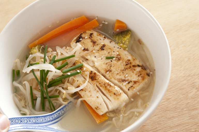 Spoon being placed in delicious Asian cuisine meal of soup broth with milkfish chunks, seasoning, chives, carrots and noodles