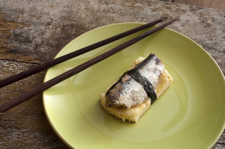 Pair of dark brown chopsticks on round green plate of omelete or tamagoyaki and fish bound together with seaweed over rustic wooden table