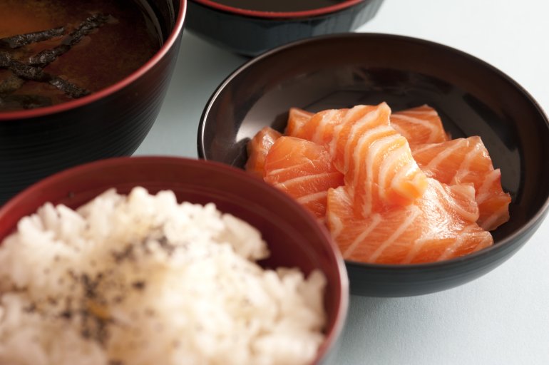 Selective focus view on round bowl of raw red salmon chunks next to seasoned white rice and other dishes