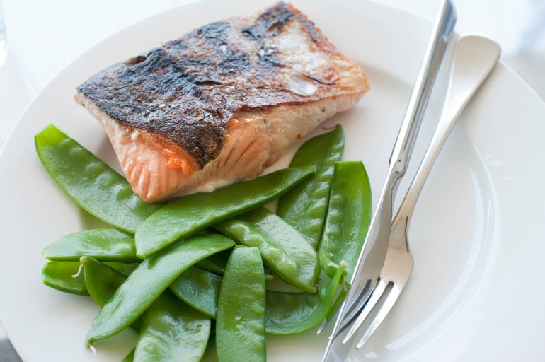 Cooked salmon fillet steak with sugarsnap peas served on a plain white plate with silver cutlery