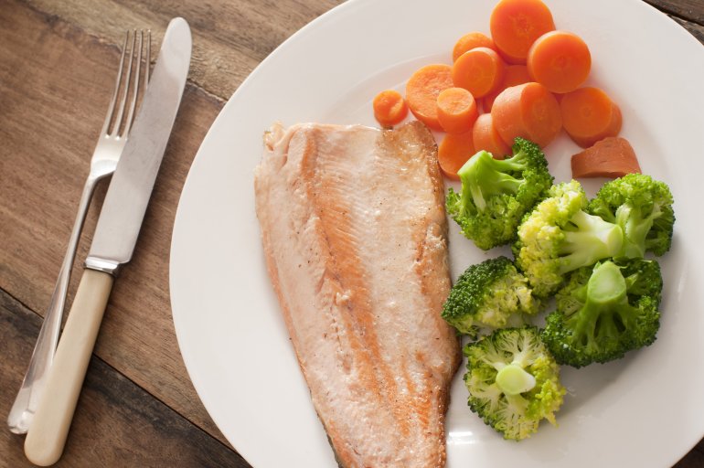 First person perspective view over single round plate of cooked trout dinner with green broccoli and slices of carrots next to fork and knife over wooden table