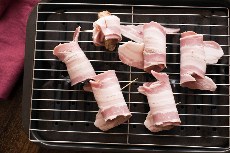 Making a batch of pigs in blankets or bacon rolls around pork sausages laid on a grill pan ready to place in the oven viewed from above