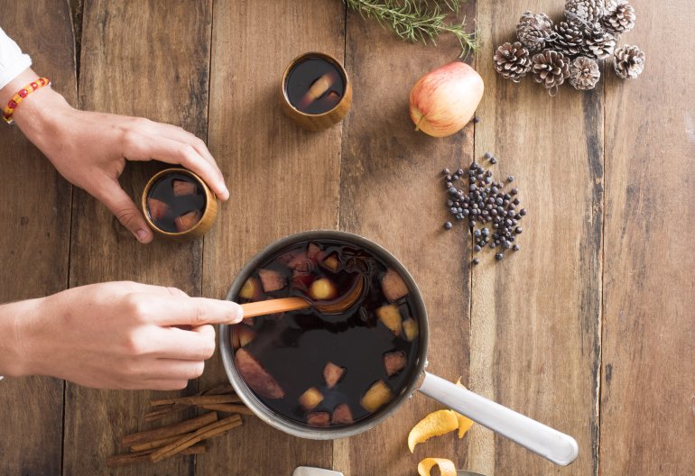 Person serving fresh hot mulled red wine with fruit and spices from a large pot into mugs using a ladle, overhead view with ingredients