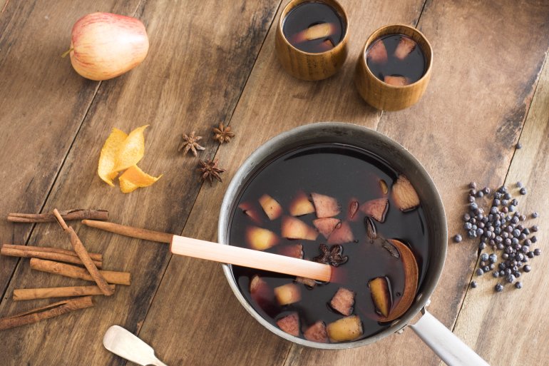 Pot of freshly made hot spicy mulled red wine surrounded by spice ingredients and fruit on a wooden table viewed from overhead