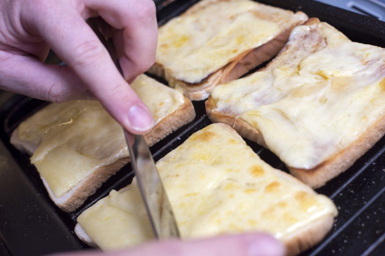 Pair of hands from unidentifiable person preparing four square slices of bread grilled with melted cheese