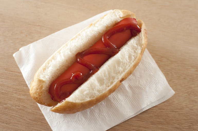 First person view on thick hot dog in white bread bun with stream of red ketchup applied to the top over napkin on table