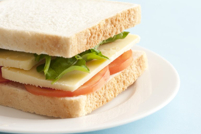Close up on side of sandwich with thick slices of yellow cheese, green lettuce leaves and round tomato slices on plate over blue