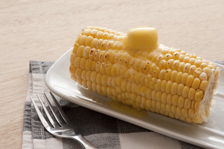 Buttery tasty grilled sweet corn on the cob with a large blob of butter on top melting and running down the kernels, served on a plate