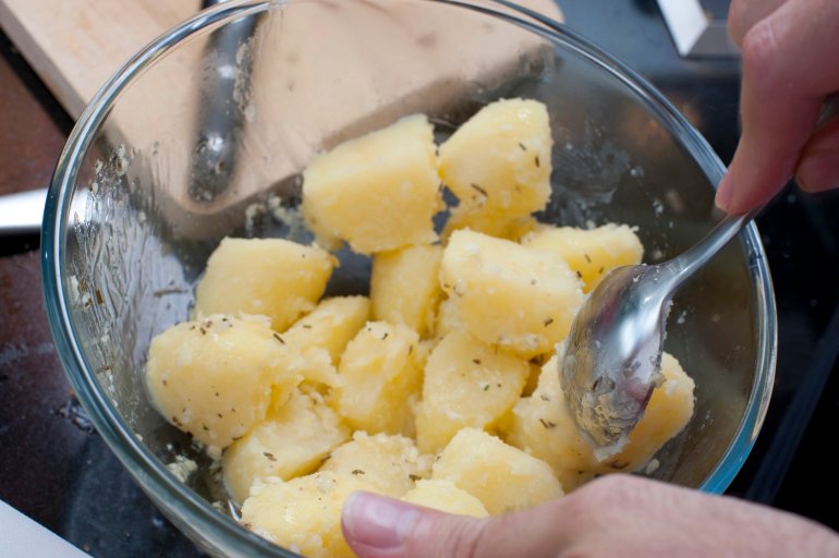 Diced boiled potatoes served in a glass bowl with a hand holding a spoon ready to serve them as a vegetable accompaniment to a meal